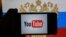 A hand holding a smartphone with YouTube's logo on the screen, in front of a computer screen in St. Petersburg, Russia showing the Russian national flag and coat of arms